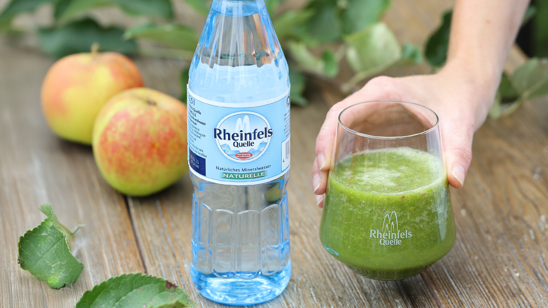 Auf einen Tisch im Garten wird ein glas mit dem grünen Smoothie abgestellt. Daneben eine Flasche Rheinfels Quelle Naturelle 0,5 L Pet Mehrweg.
