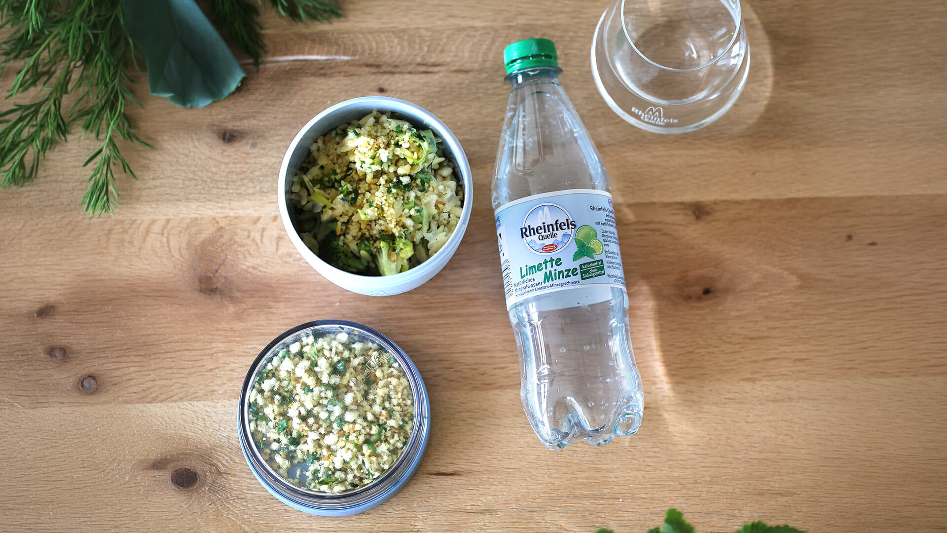 One-Pot Orzo mit Zitronen-Kräuter-Crunch mit einer Flasche Rheinfels Quelle Limette-Minze 0,5 Liter PET-Mehrweg. Beides auf einem Holzschneidebrett dekoriert mit Dill, Brokkoli und Petersilie