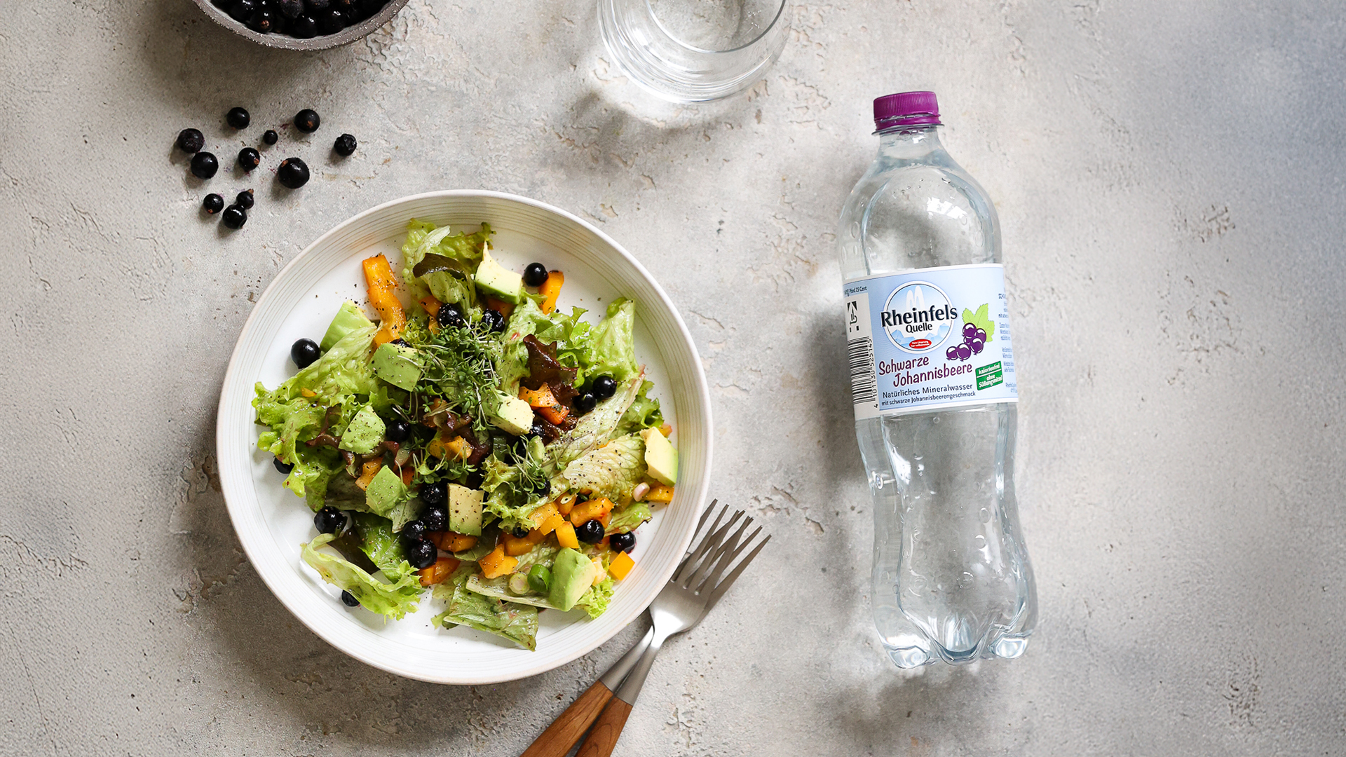 Ein gemischter Salat mit Johannisbeeren steht auf der Arbeitsplatte und eine Flasche Rheinfels Quelle Schwarze Johannisbeere liegt rechts daneben.