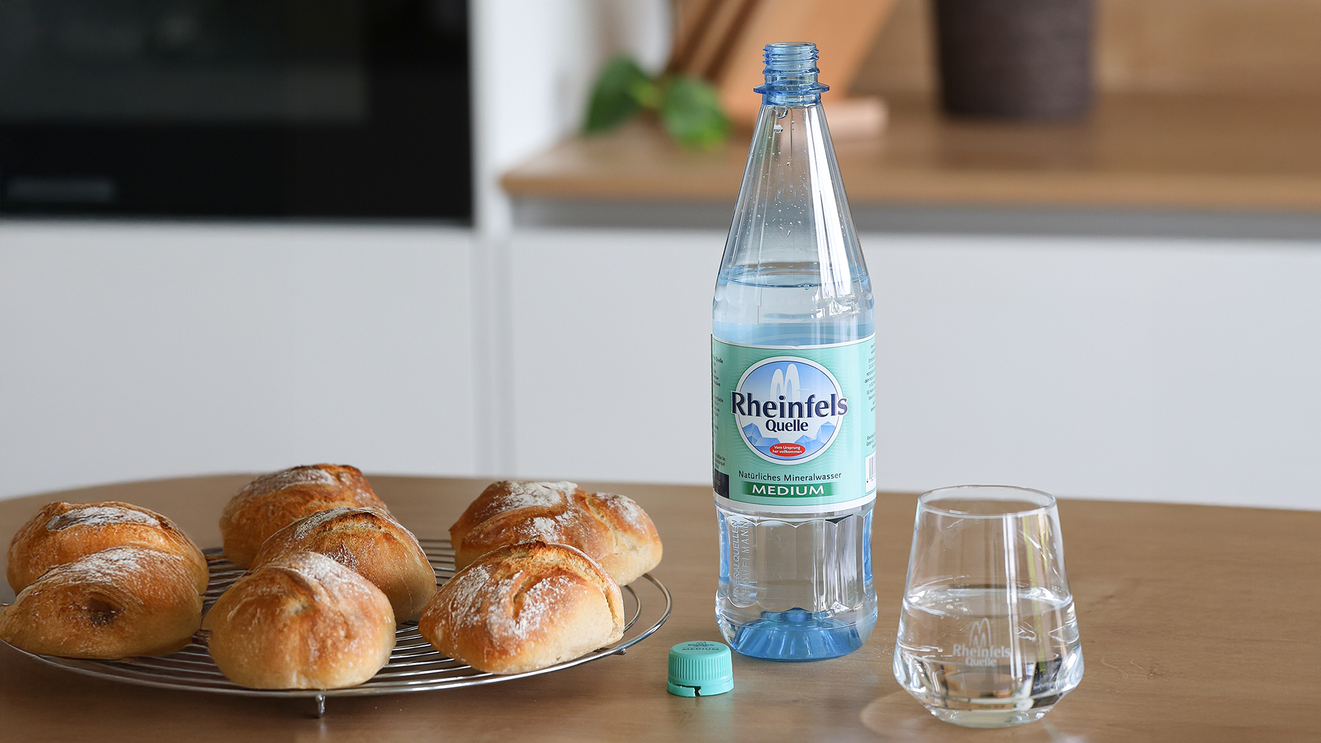 Selbstgebackene Dinkel-Sauerteigbrötchen liegen zum Auskühlen auf einem Gitter, daneben eine geöffnete Flasche Rheinfels Quelle 1 Liter PET Mehrweg Medium.