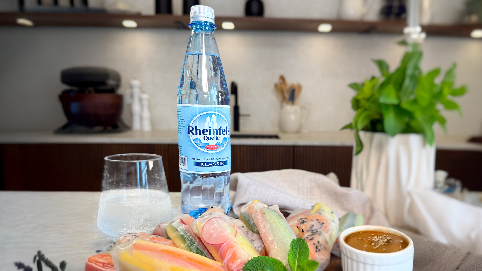 Sommerrollen mit Erdnusssoße angerichtet auf einem Holzteller. Dazu eine Flasche Rheinfels Quelle Klassik