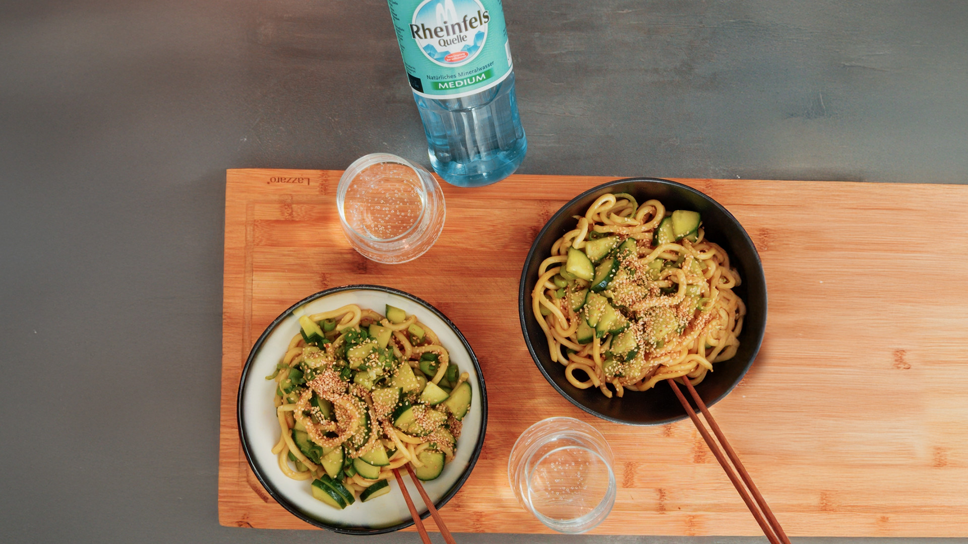 Spicy Udon mit Gurke und Sesam mit einer Flasche Rheinfels Quelle Medium auf einem Holzbrett in Schüssel serviert