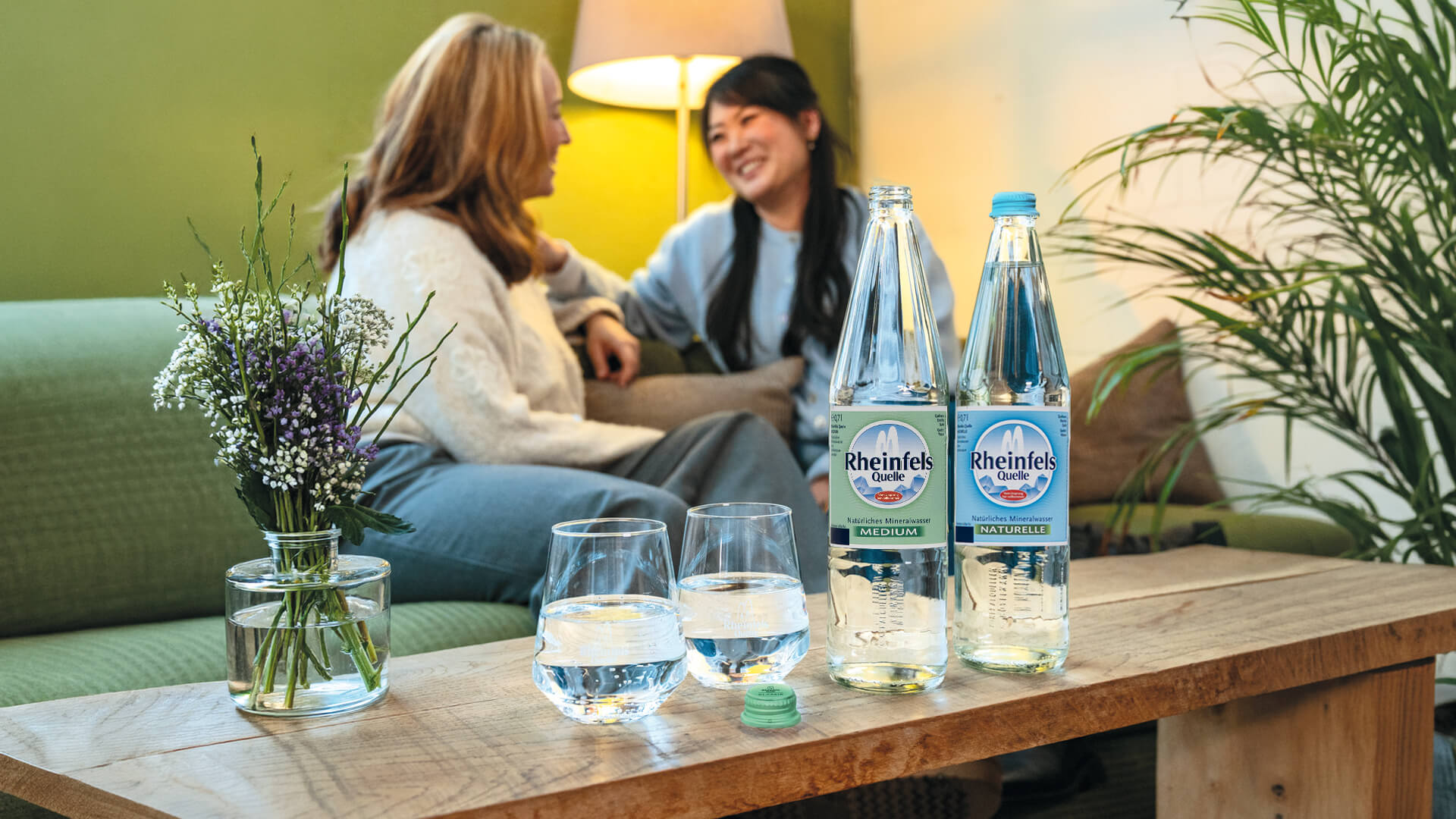 Zwei Frauen auf einem Sofa, im Vordergrund zwei gefüllte Gläser mit Rheinfels Quelle Klassik und Naturelle Flaschen auf dem Tisch