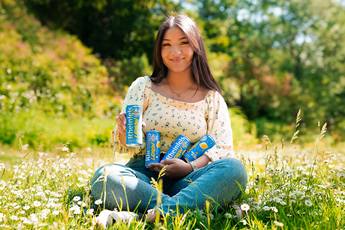 Junge Frau auf einer Wiese sitzend mit mehreren Rheinfels Quelle Lemon Dosen in der Hand und auf dem Arm 