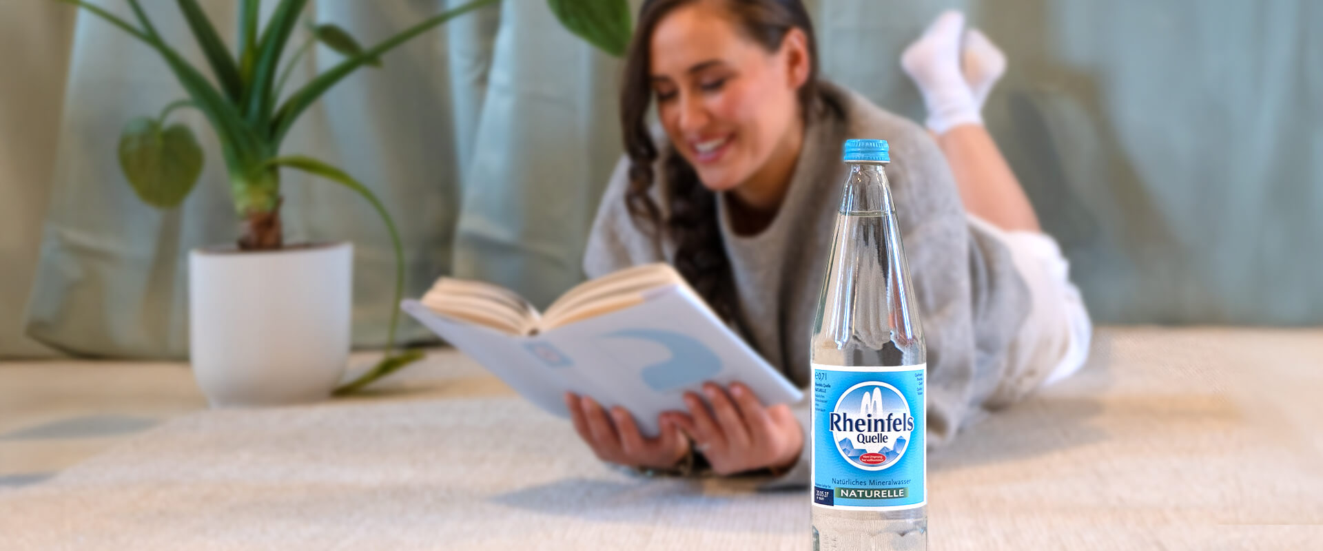 Eine Frau liegt in ihrem Wohnzimmer auf dem Fußboden und liest ein Buch. Eine Flasche Rheinfels Quelle Naturelle 0,7 Liter Glas steht auf dem Teppich.