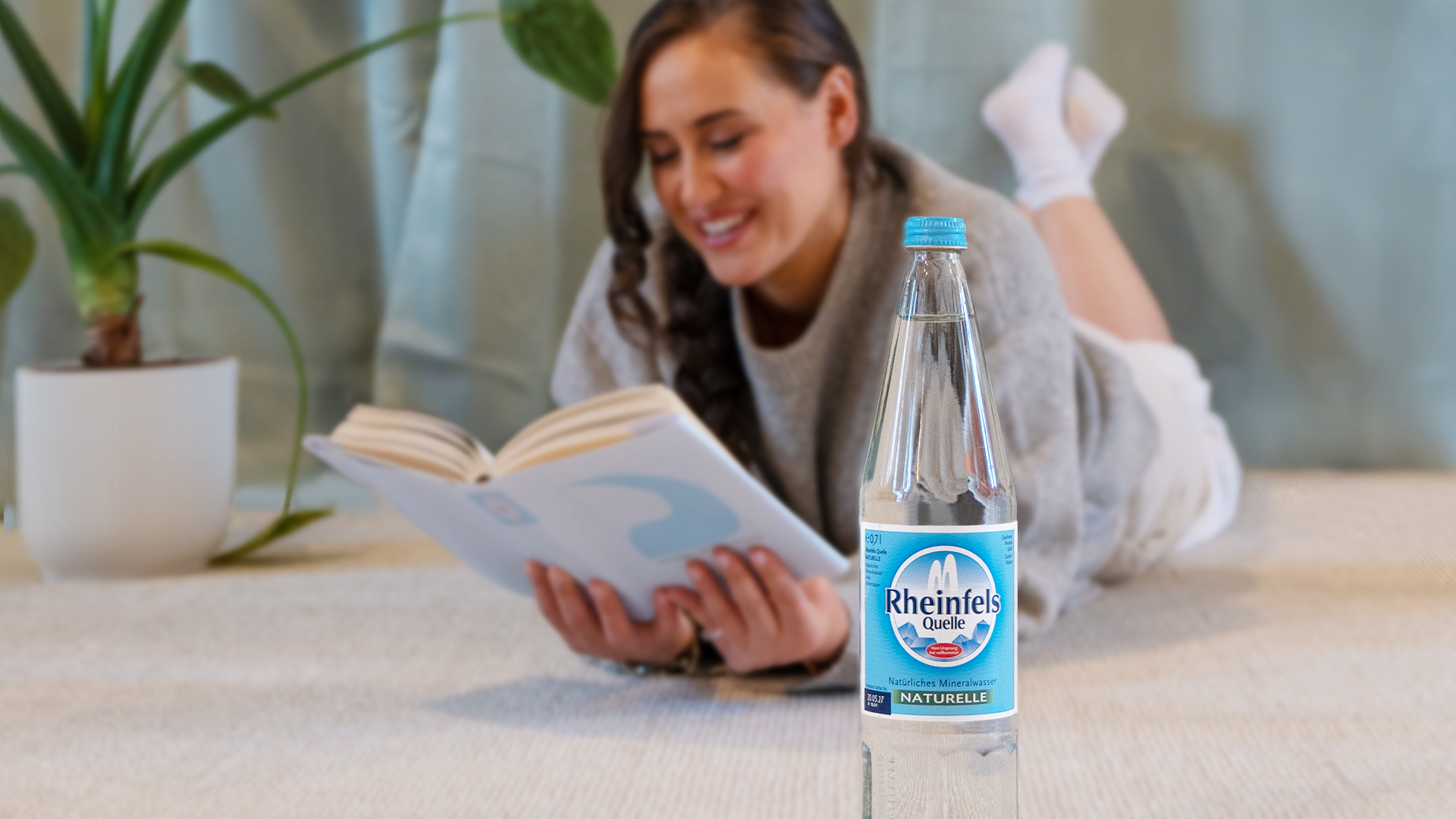 Eine Frau liegt auf einem Teppich und liest, eine Glasflasche Rheinfels Quelle Naturelle steht neben ihr.